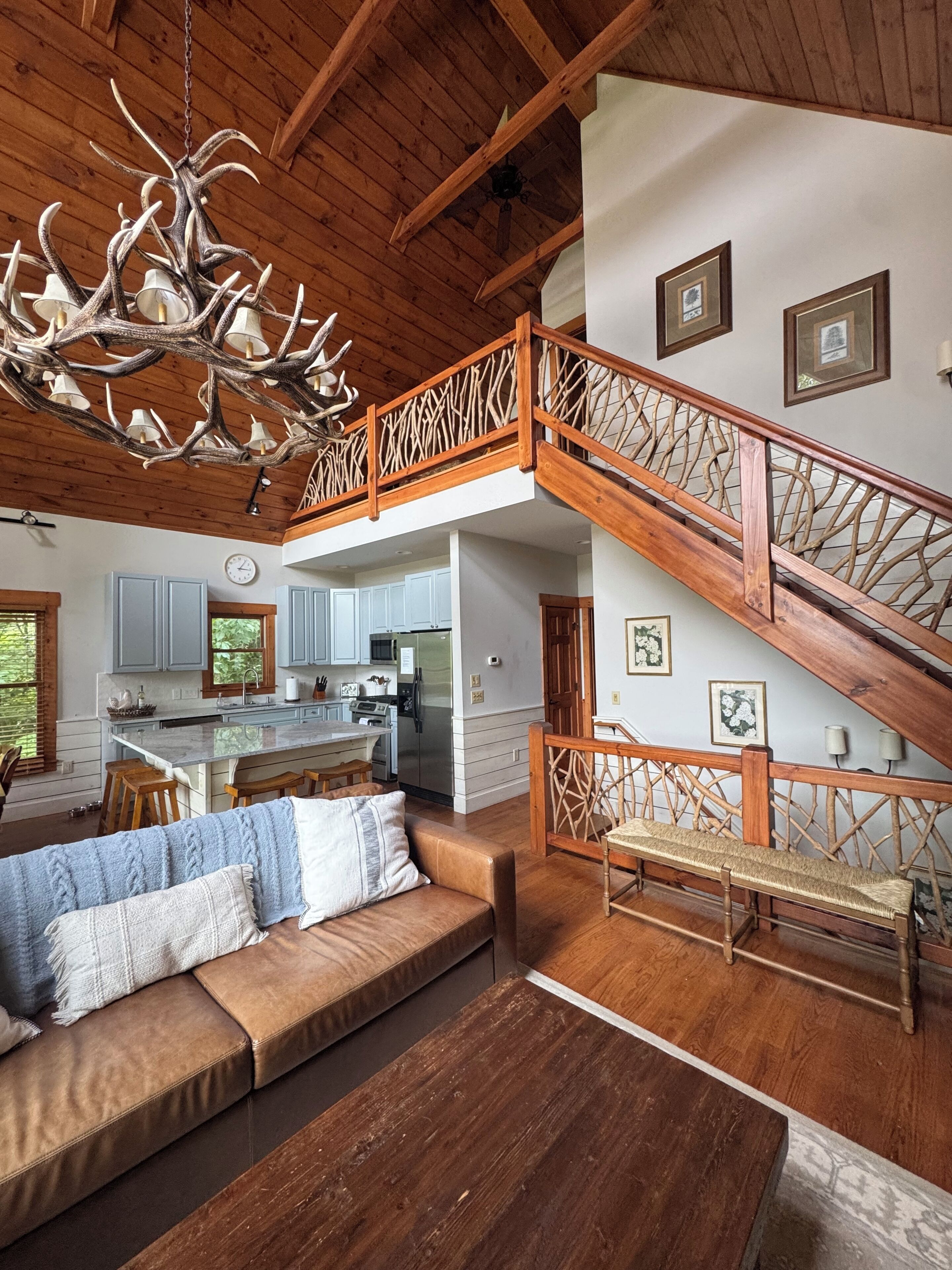 A spacious living area with a wooden ceiling, a large chandelier, a wooden staircase, and a kitchen in the background.