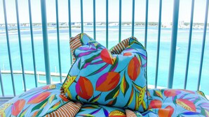 A colorful pillow with a tropical leaf pattern, a view of the ocean and distant buildings, and a railing.