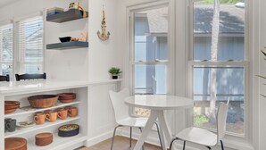 A bright room with white walls, a white table, and two white chairs. Shelves above the table hold various items, and there are large windows with blinds.