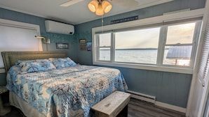 A bedroom with a bed, a wooden bench, a ceiling fan, a window with a view of the water, and a wall-mounted air conditioner. - Charming Cottage  LITERALLY on  Lake --Pet Friendly with Guest Cottage (Romulus)