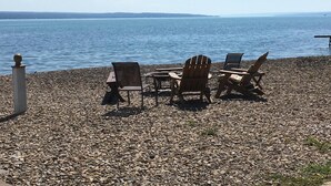 A pebbled beach with wooden Adirondack chairs and a table, overlooking a calm body of water. - Charming Cottage  LITERALLY on  Lake --Pet Friendly with Guest Cottage (Romulus)