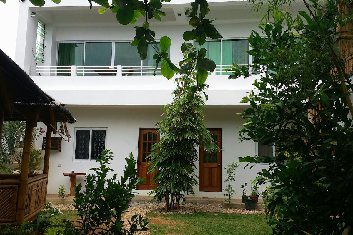 A two-story building with a balcony, a small garden area with potted plants, and a wooden structure to the left.