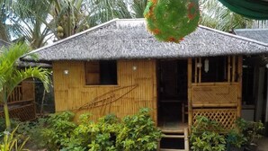 A small, single-story bamboo house with a thatched roof, surrounded by lush greenery and a concrete pathway. - HangOut Beach & Resort (Malay)