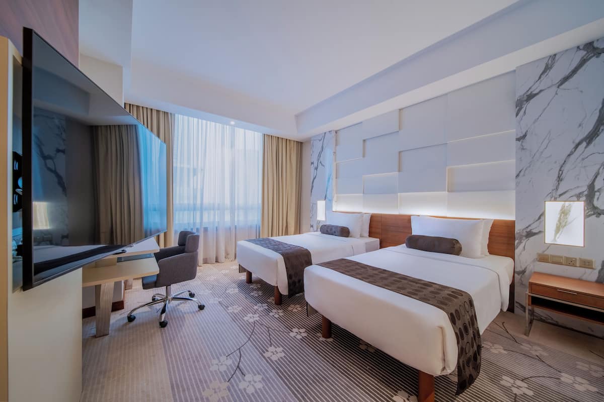 Une chambre d’hôtel avec deux lits, un bureau avec une chaise, une télévision et une grande fenêtre avec des rideaux.