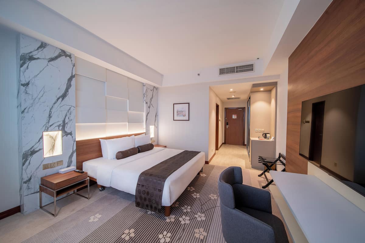 Une chambre d’hôtel moderne, dotée d’un grand lit, d’un bureau, d’une chaise, d’une télévision et d’une salle de bain visible à travers une porte ouverte.