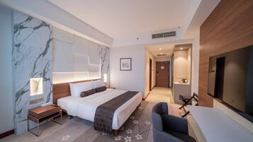 Habitación de hotel moderna con una cama grande, un escritorio, una silla, un televisor y un baño visible a través de una puerta abierta.