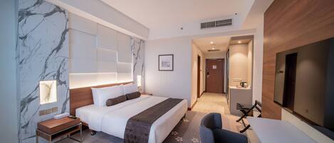 Habitación de hotel moderna con una cama grande, un escritorio, una silla, un televisor y un baño visible a través de una puerta abierta.