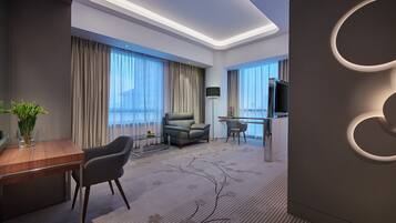 A modern hotel room with a dining area, a sofa, a TV, and large windows with curtains.