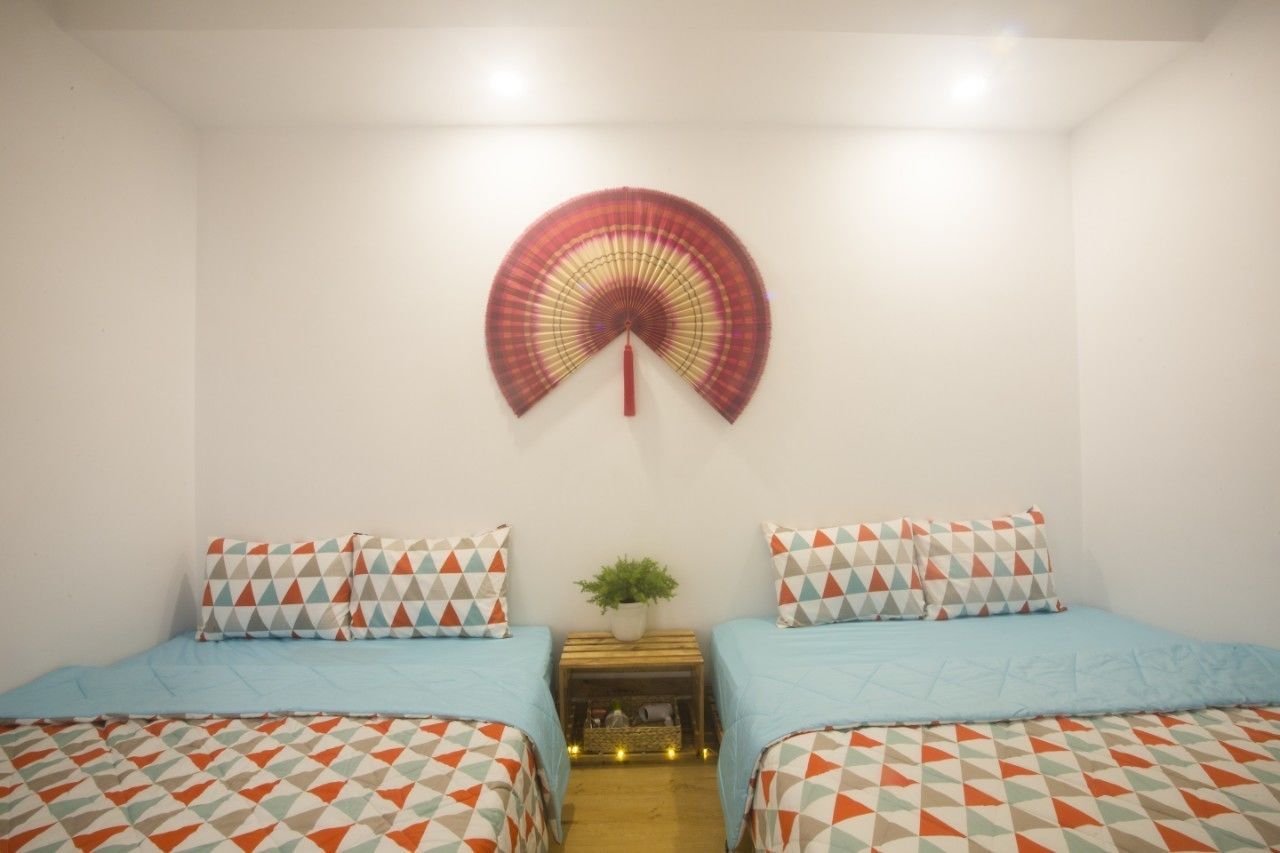 A room with two beds, each with patterned bedding, a wall-mounted fan, and a small plant on a wooden nightstand.