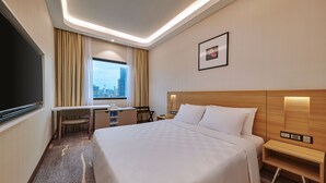 Ein Hotelzimmer mit einem großen Bett, einem Schreibtisch, einem Sessel, einem Fernseher und einem Fenster mit Blick auf die Stadt.