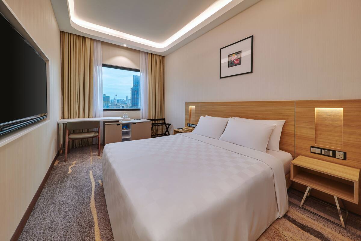 A hotel room with a large bed, a desk, a chair, a TV, and a window with a city view.