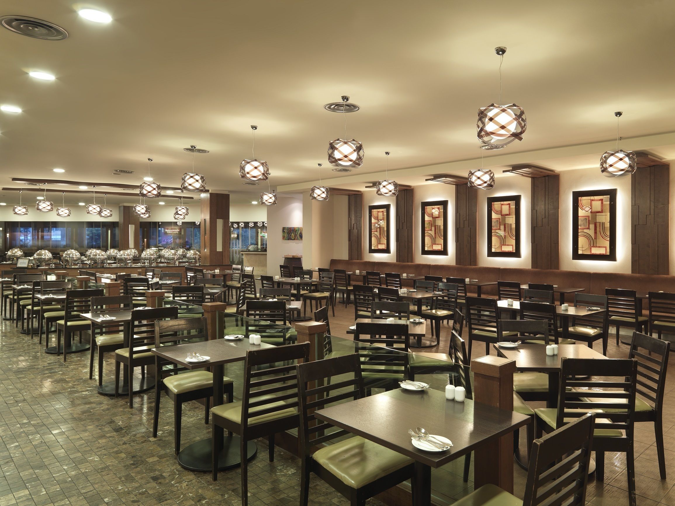 A spacious restaurant with multiple dining tables and chairs, overhead lighting fixtures, and large windows in the background.