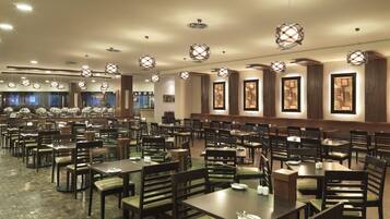 A spacious restaurant with multiple dining tables and chairs, overhead lighting fixtures, and large windows in the background.