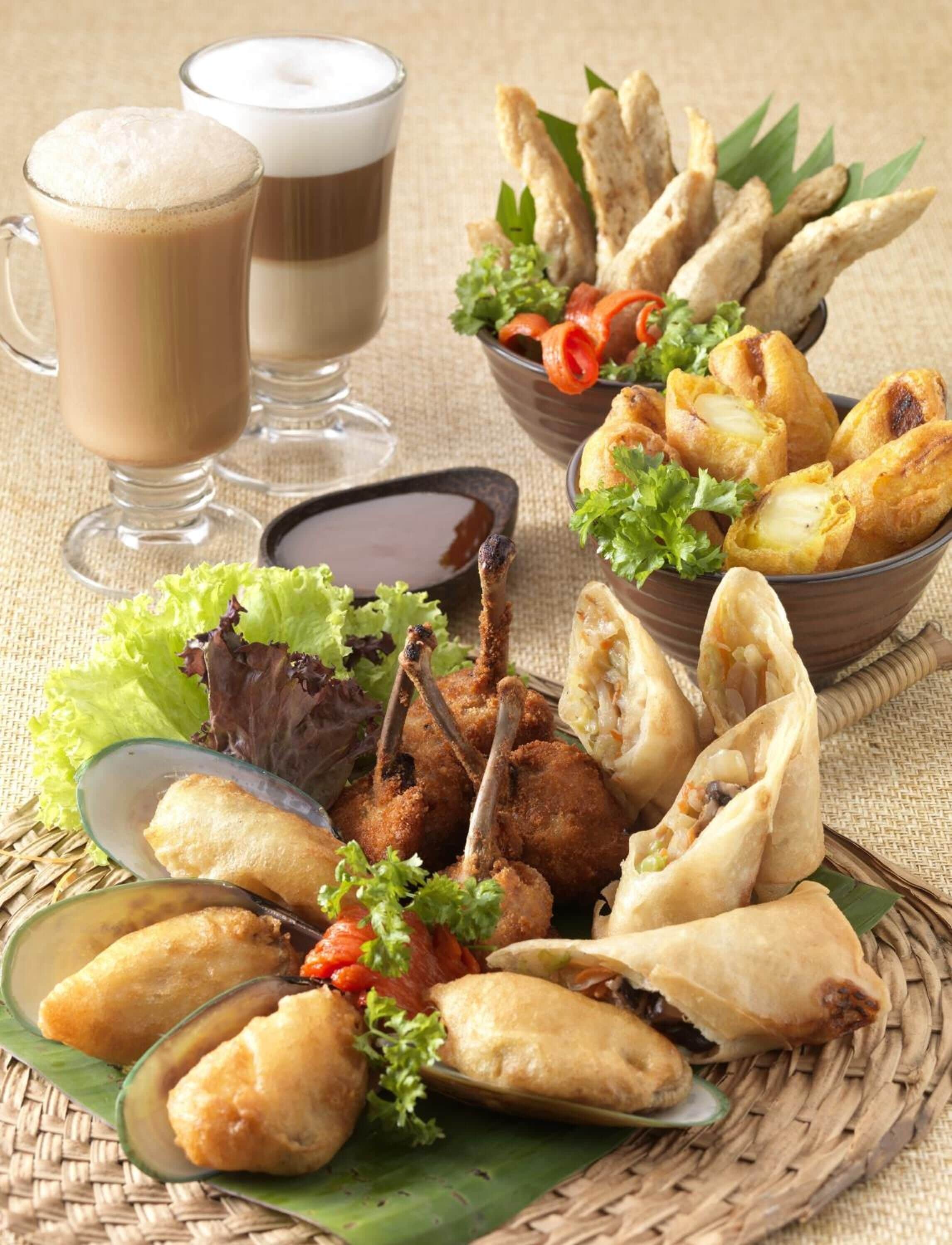 Diversos pratos fritos, incluindo samosas, frango frito e uma salada, acompanhados por dois copos de uma bebida castanha.
