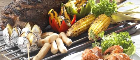 Variedad de carnes y verduras a la parrilla sobre una mesa de madera, incluyendo un pollo asado grande, brochetas y mazorcas de maíz.
