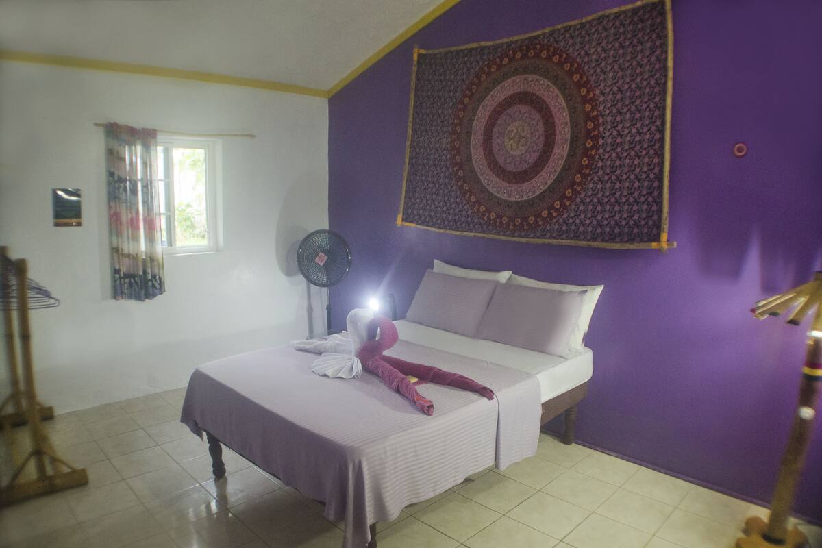 Una habitación con cama, un tapiz, una ventana con cortinas y un ventilador.