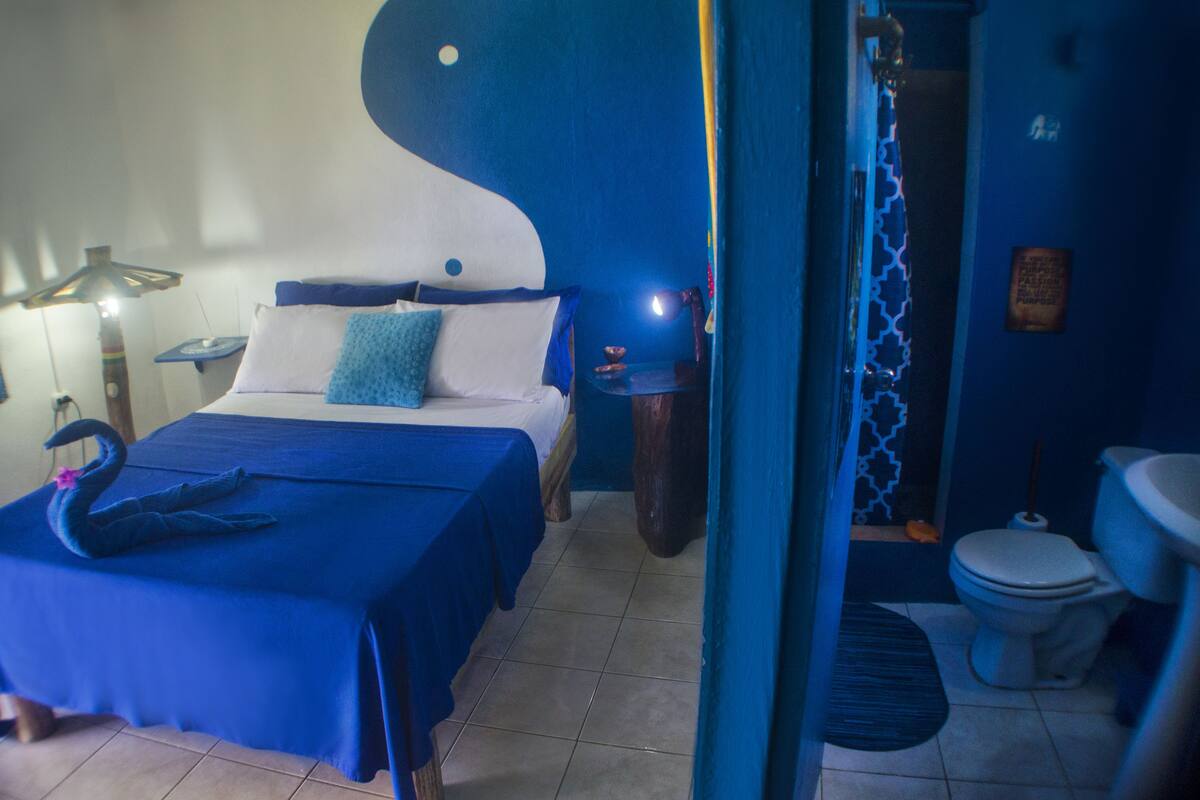 Una habitación con cama, mesita de noche, lámpara, adorno con forma de cisne y un baño con inodoro y lavabo.