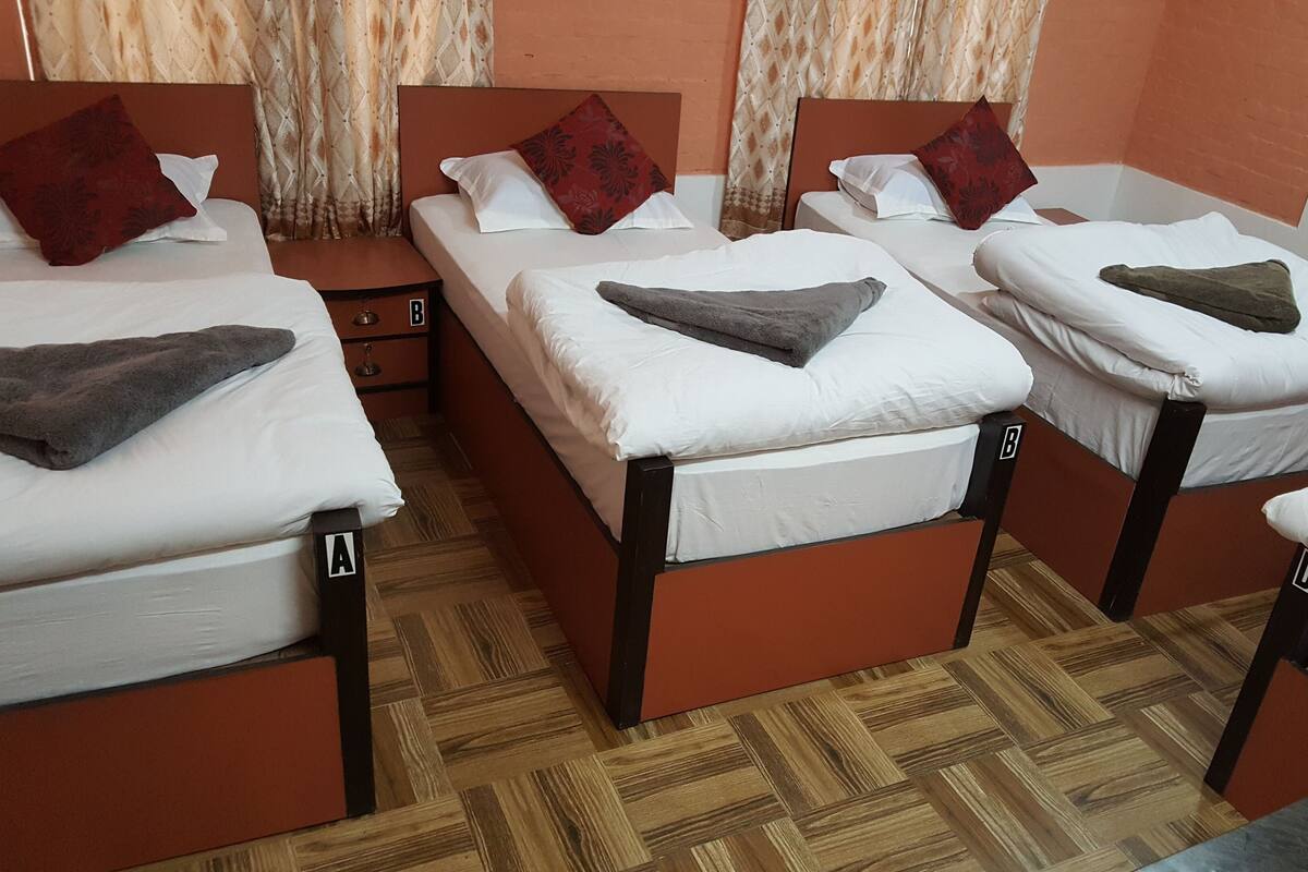 Une chambre d’hôtel avec quatre lits simples, chacun étant équipé d’un oreiller et d’une couverture. Les lits sont identifiés par les lettres A, B, C et D. La chambre est dotée d’un parquet en bois et de rideaux de couleur beige.