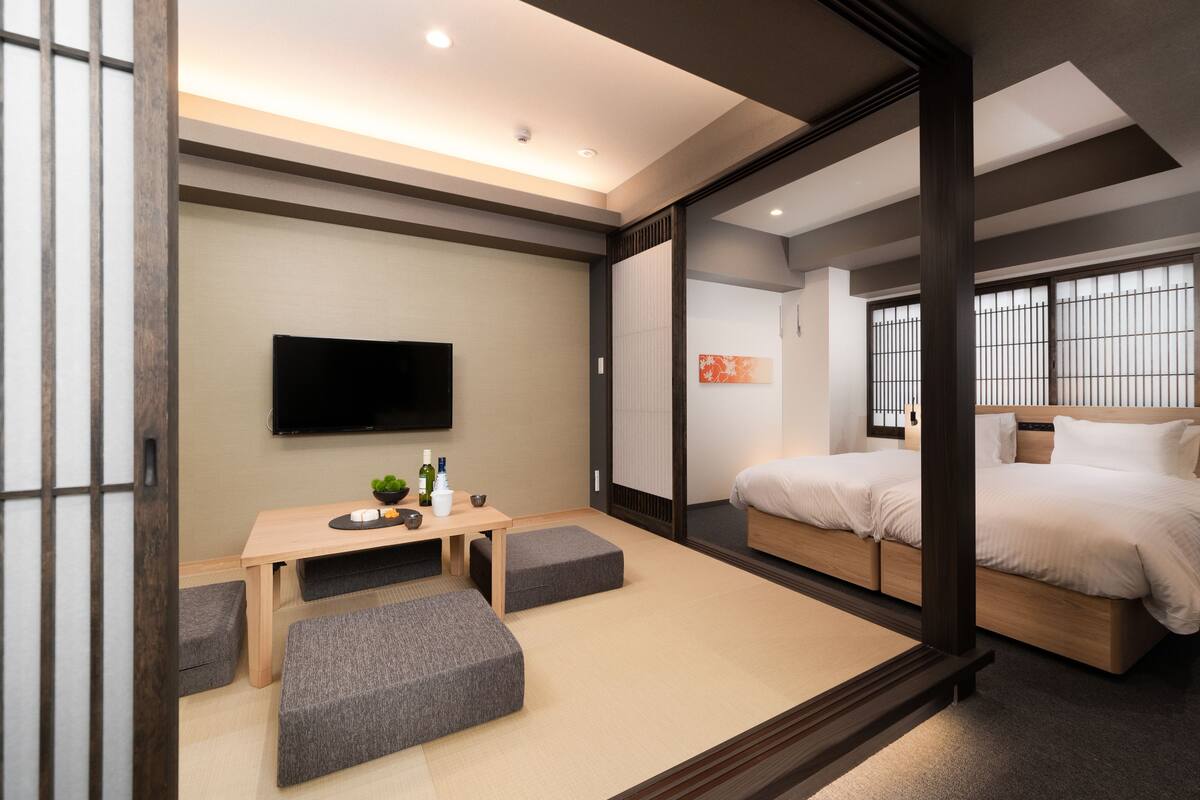 Una habitación de hotel moderna con una cama de madera, un televisor de pantalla plana, un pequeño escritorio con una planta y un divisor japonés corredero.