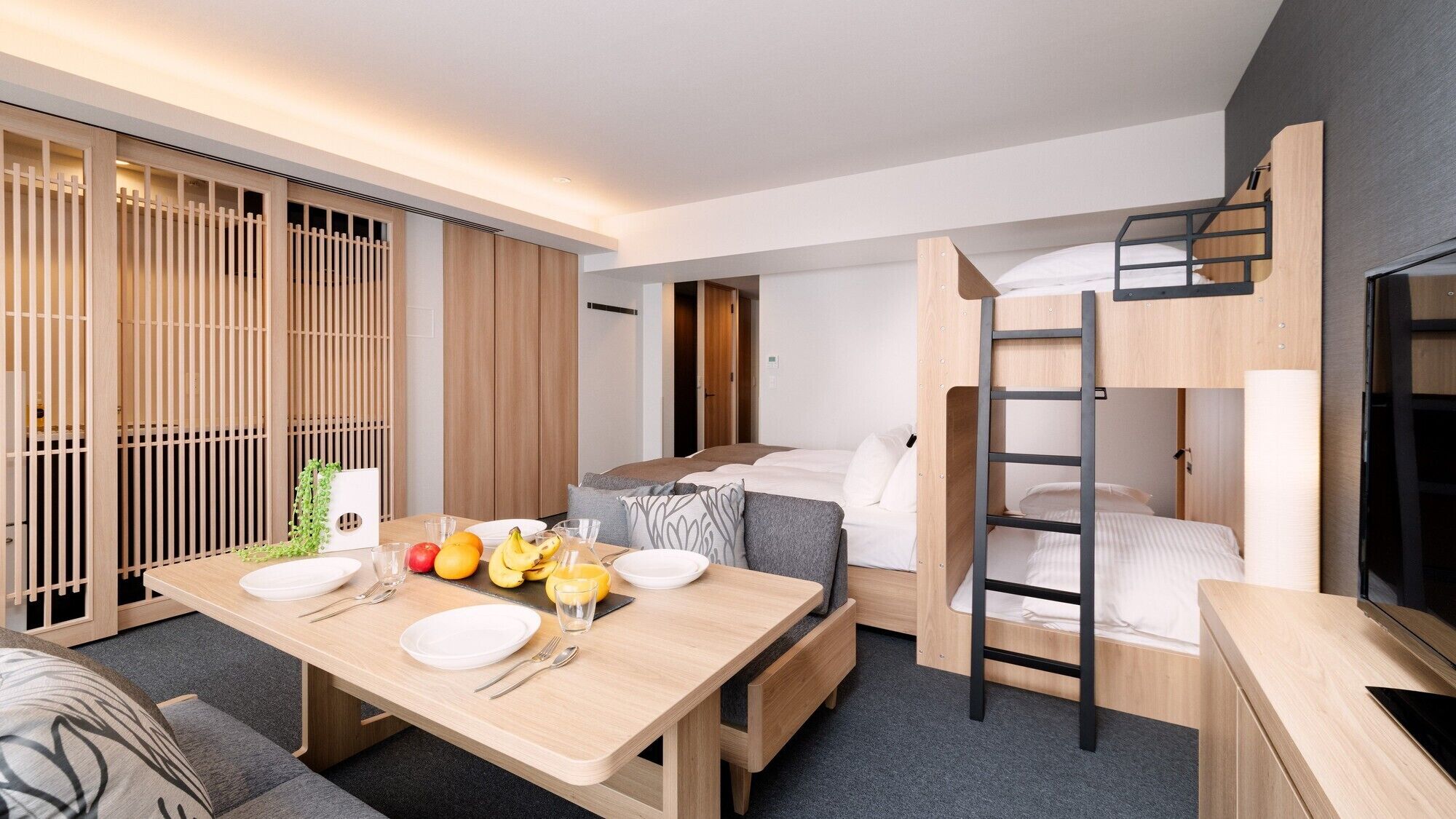 A compact hotel room with a bunk bed, a dining table with fruit and cutlery, and a sliding door with a wooden screen.