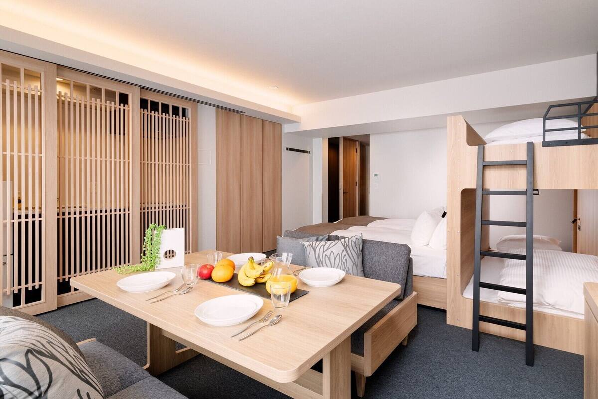 Habitación de hotel pequeña con una cama litera, una mesa de comedor con fruta y cubiertos, y una puerta corrediza con un panel de madera.