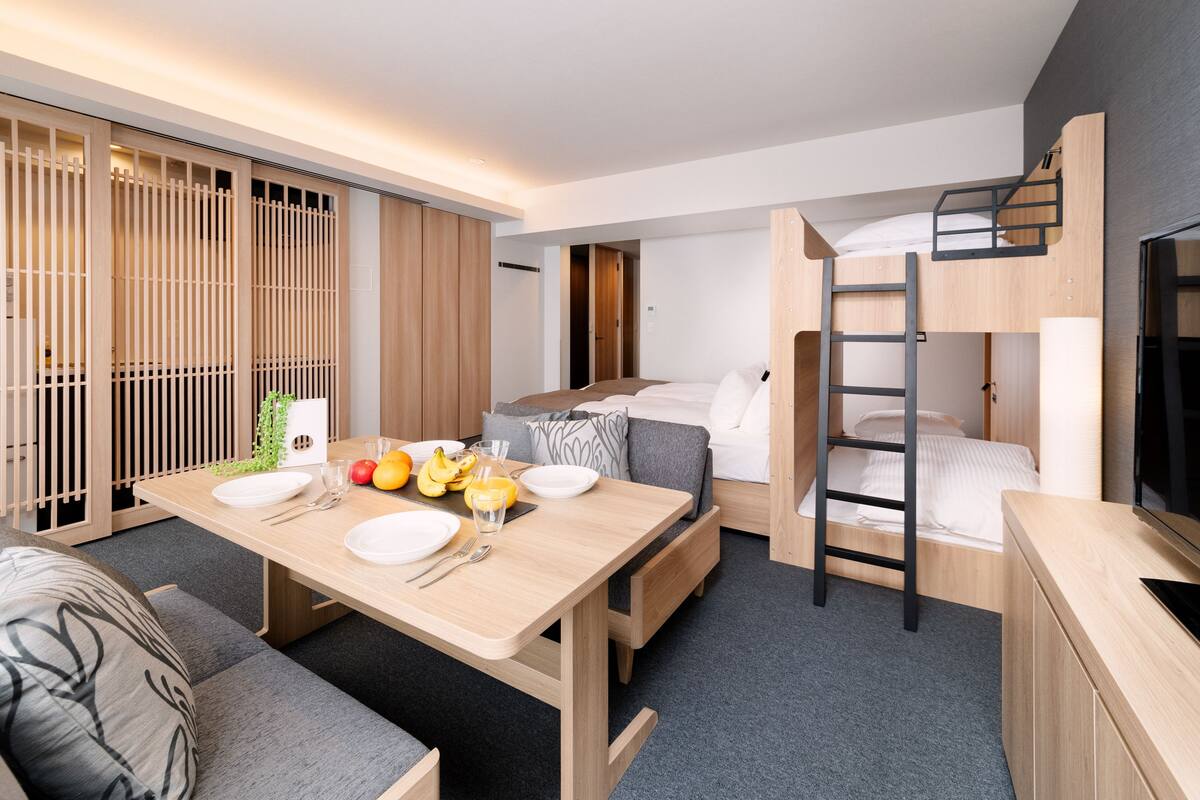 Una habitación de hotel moderna con una mesa de comedor de madera preparada para dos personas, un sofá, una cama nido con escalera y un televisor sobre un mueble de madera.