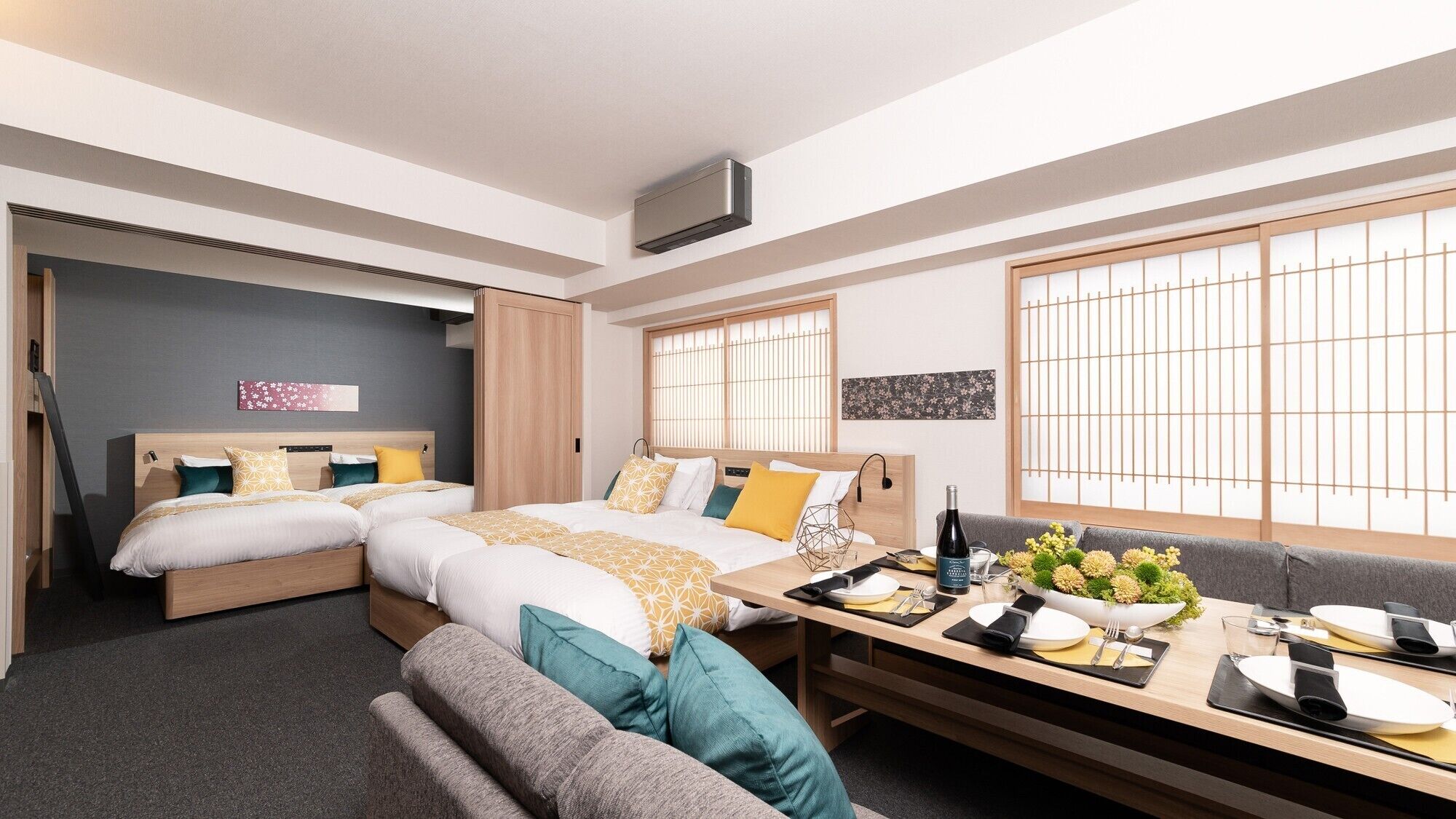 Una habitación de hotel moderna con dos camas, una mesa de comedor preparada para dos personas y ventanas amplias con vistas.