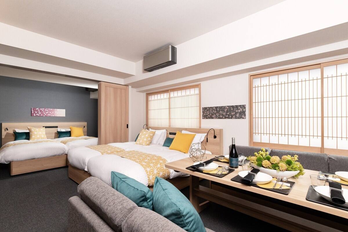 Una habitación de hotel moderna con dos camas, una mesa de comedor preparada para dos personas y ventanas amplias con vistas.