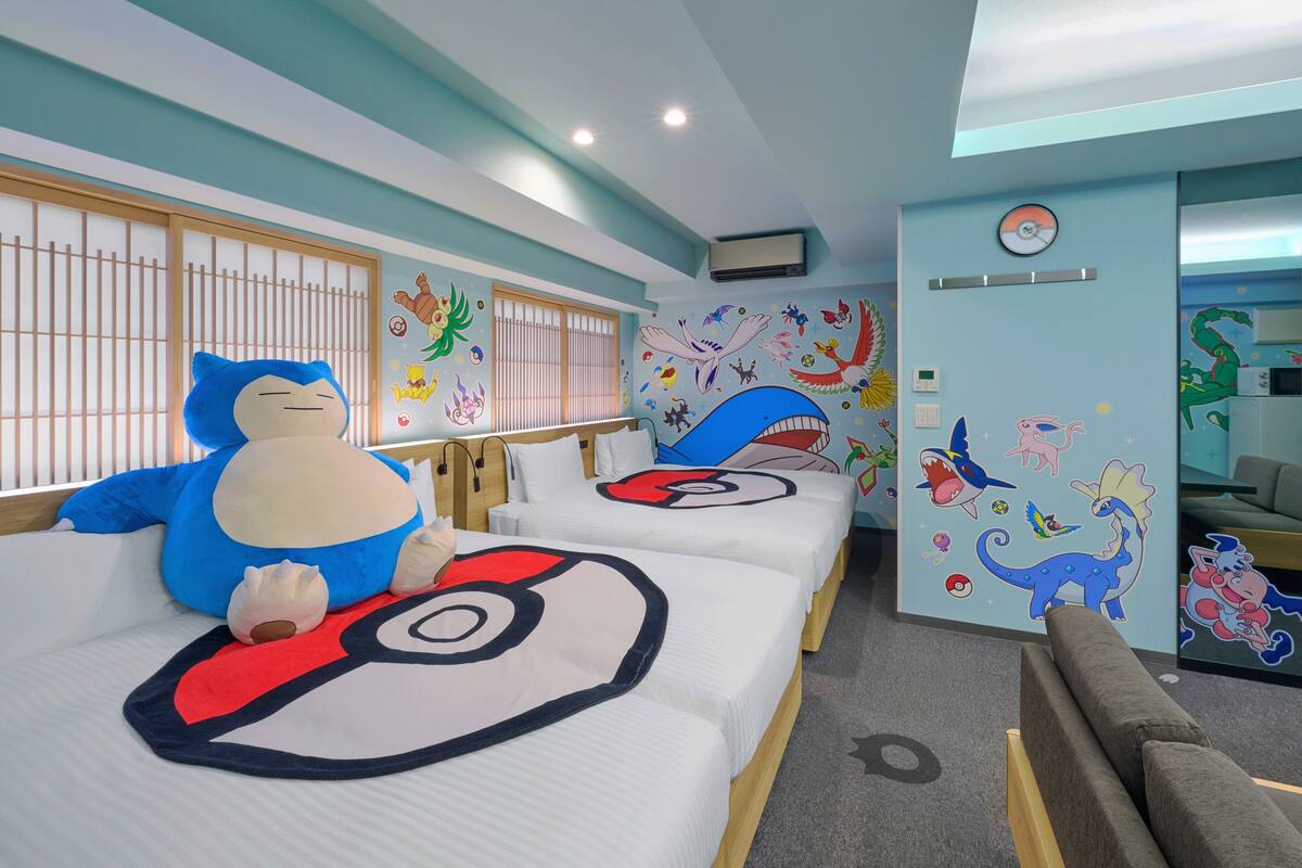 Habitación de hotel con dos camas, cada una con un almohadón de gran tamaño con un personaje de dibujos animados, una pared decorada con pegatinas de colores y un equipo de aire acondicionado visible.