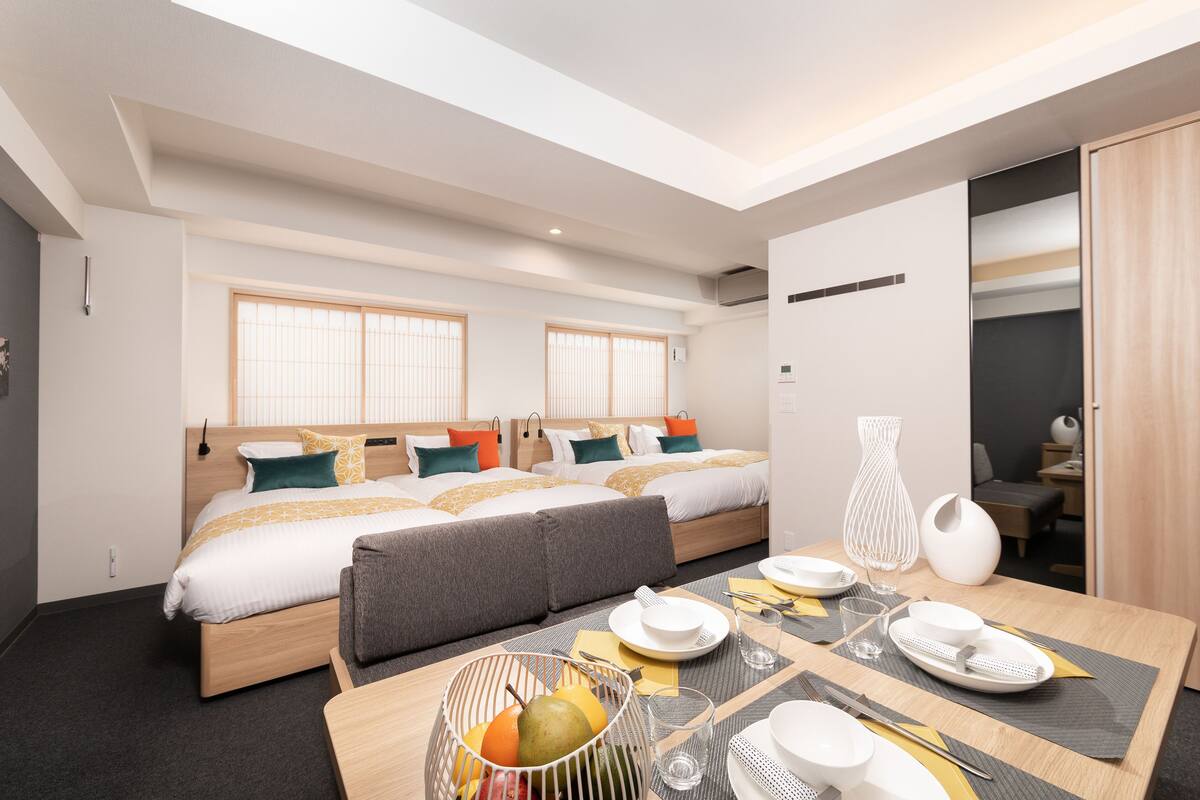 Una habitación de hotel moderna con una cama grande, una mesa de comedor con platos y vasos, y una pequeña cesta de fruta.