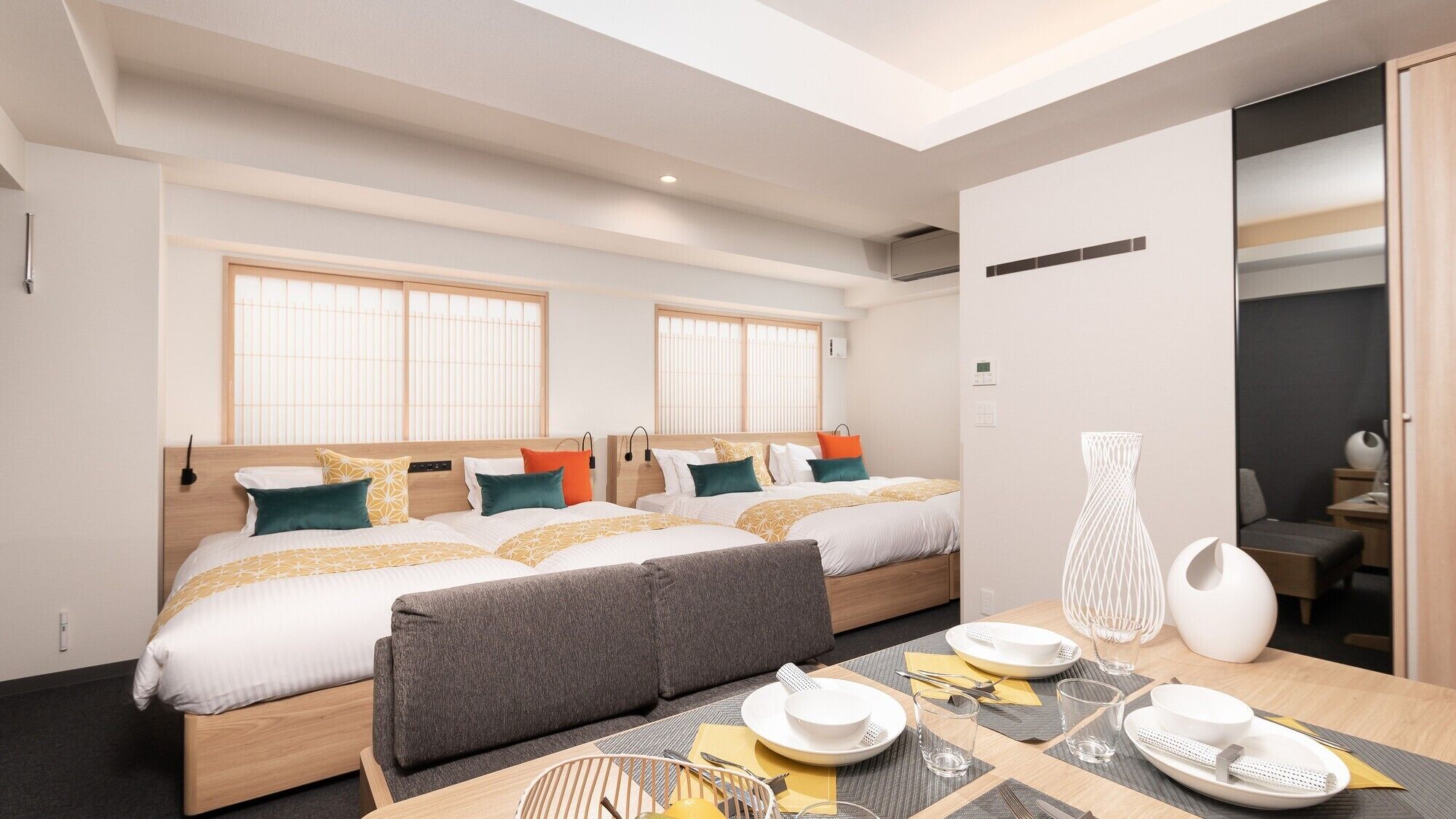 A modern hotel room with two beds, a dining area, and a view of the city.