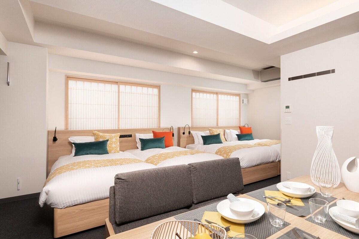 A modern hotel room with two beds, a dining area, and a view of the city.