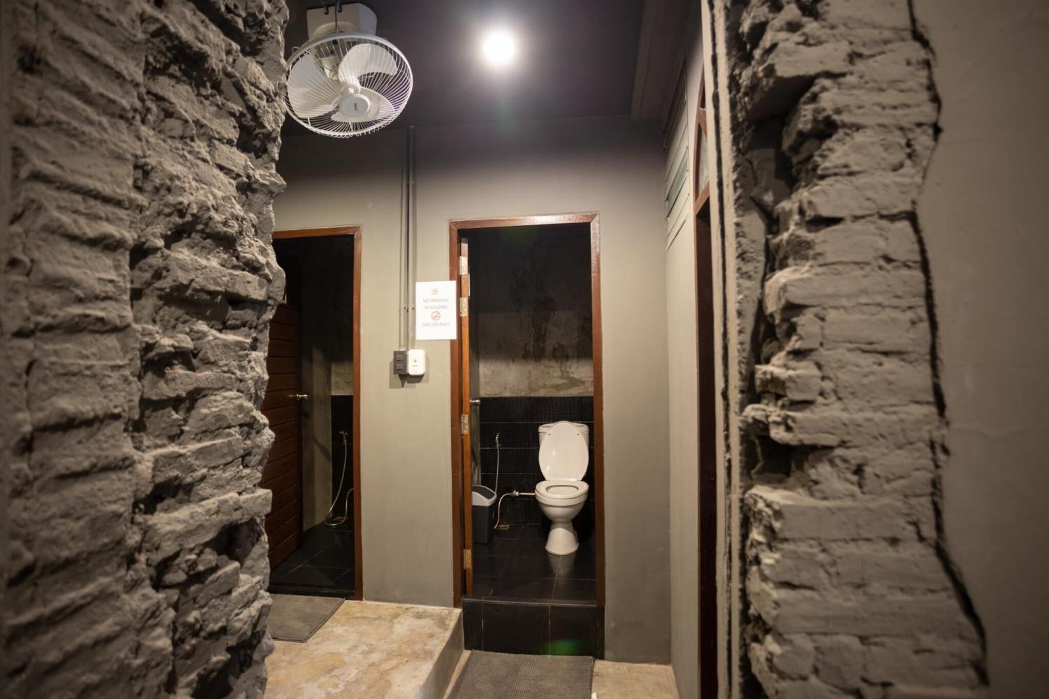 A narrow hallway with a stone wall on one side and a wooden door on the other. A ceiling fan is visible above the door.