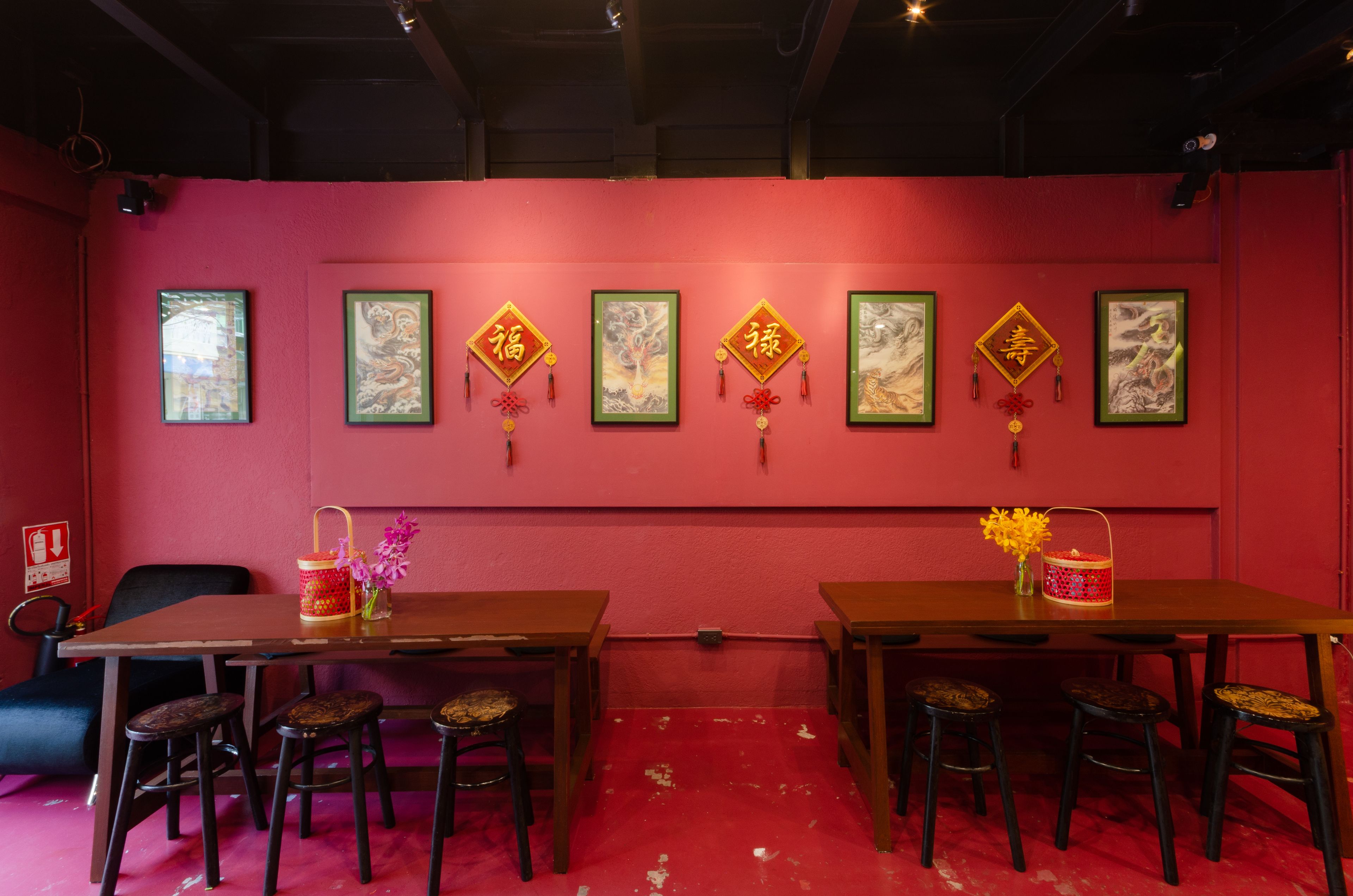 Ein Zimmer mit roter Wand, Holztischen und -stühlen. An der Wand befinden sich gerahmte Kunstwerke und ein kleiner Tisch mit Blumen in einer Vase.