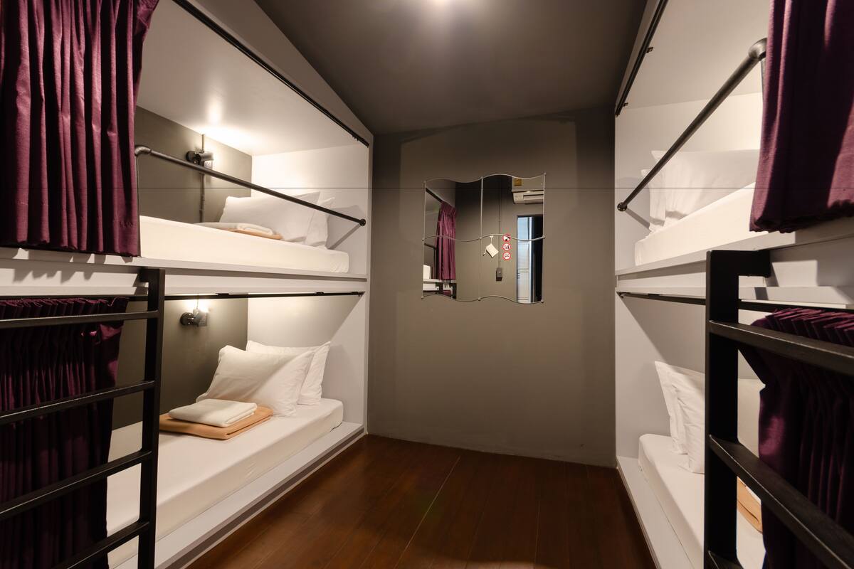 A narrow, dimly lit corridor with bunk beds on either side, a mirror on the wall, and a wooden floor.