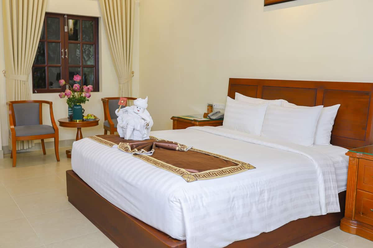 A neatly made bed with white linens, a wooden headboard, and a decorative throw. There is a wooden nightstand with a lamp and a small table with a vase of flowers. A chair is positioned near the window.
