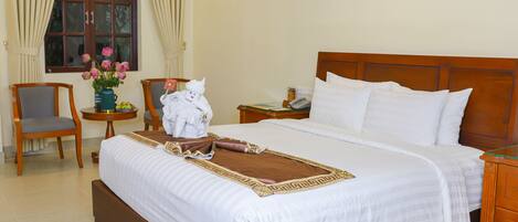 A neatly made bed with white linens, a wooden headboard, and a decorative throw. There is a wooden nightstand with a lamp and a small table with a vase of flowers. A chair is positioned near the window.