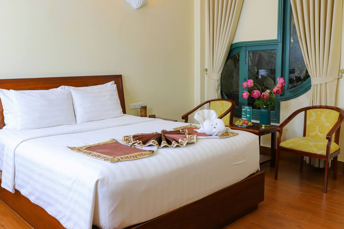 A neatly made bed with white linens, a decorative pillow, and a brown throw blanket. There is a small table with a vase of flowers and a fruit bowl. Two chairs with yellow upholstery are placed beside the bed.