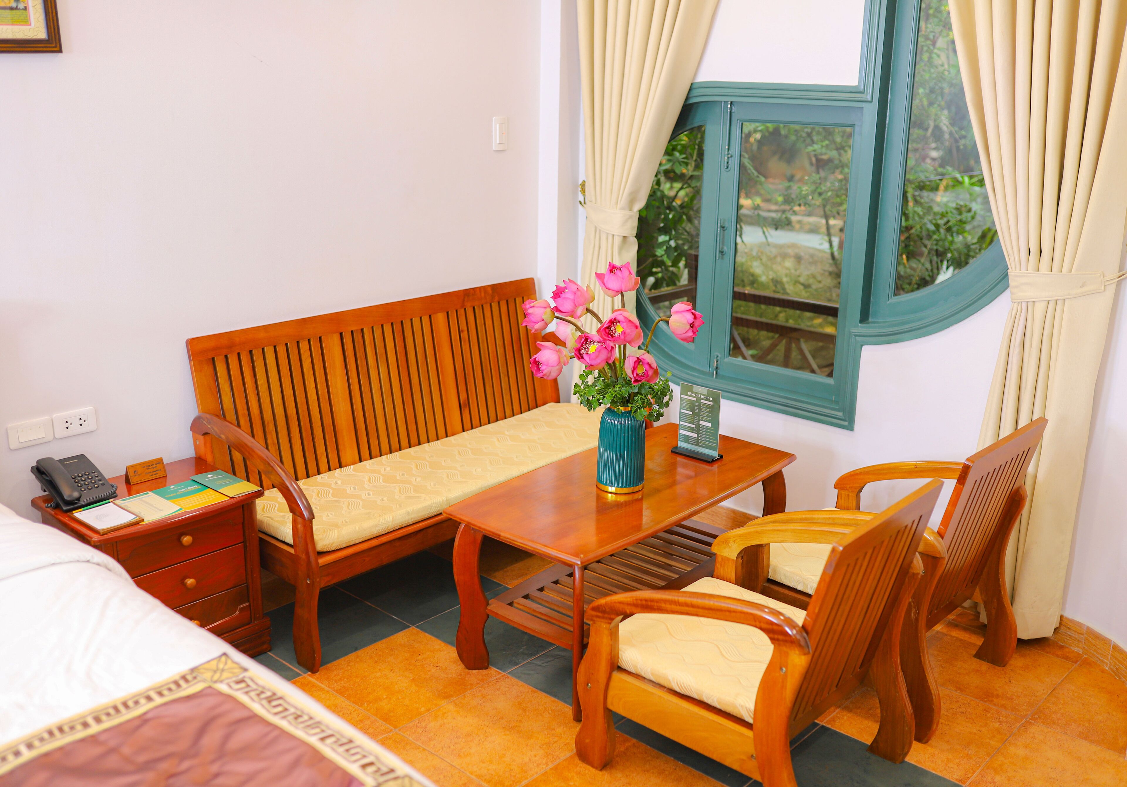 Ein Zimmer mit einem Bett, Holzmöbeln, einem Tisch mit Blumenvase und einem Vorhangfenster.