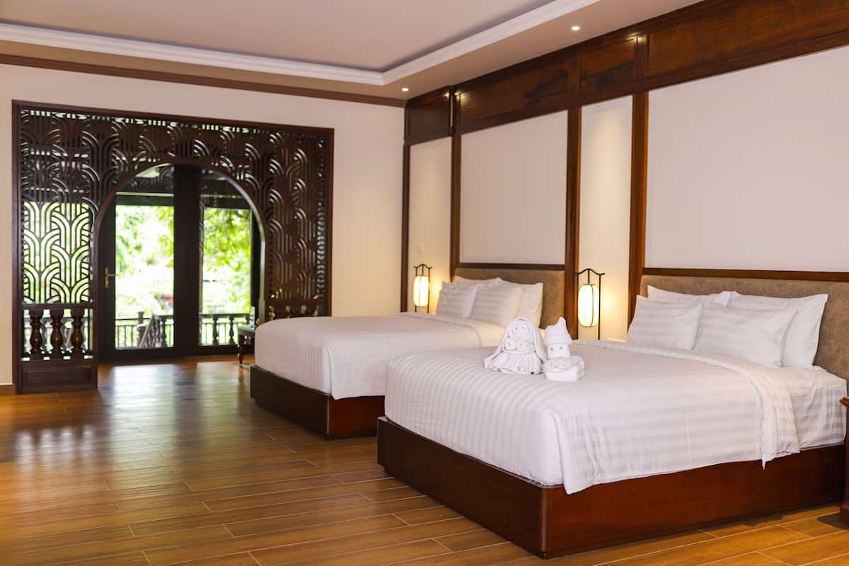 A hotel room with two beds, wooden flooring, a decorative wooden door, and a view through the door.