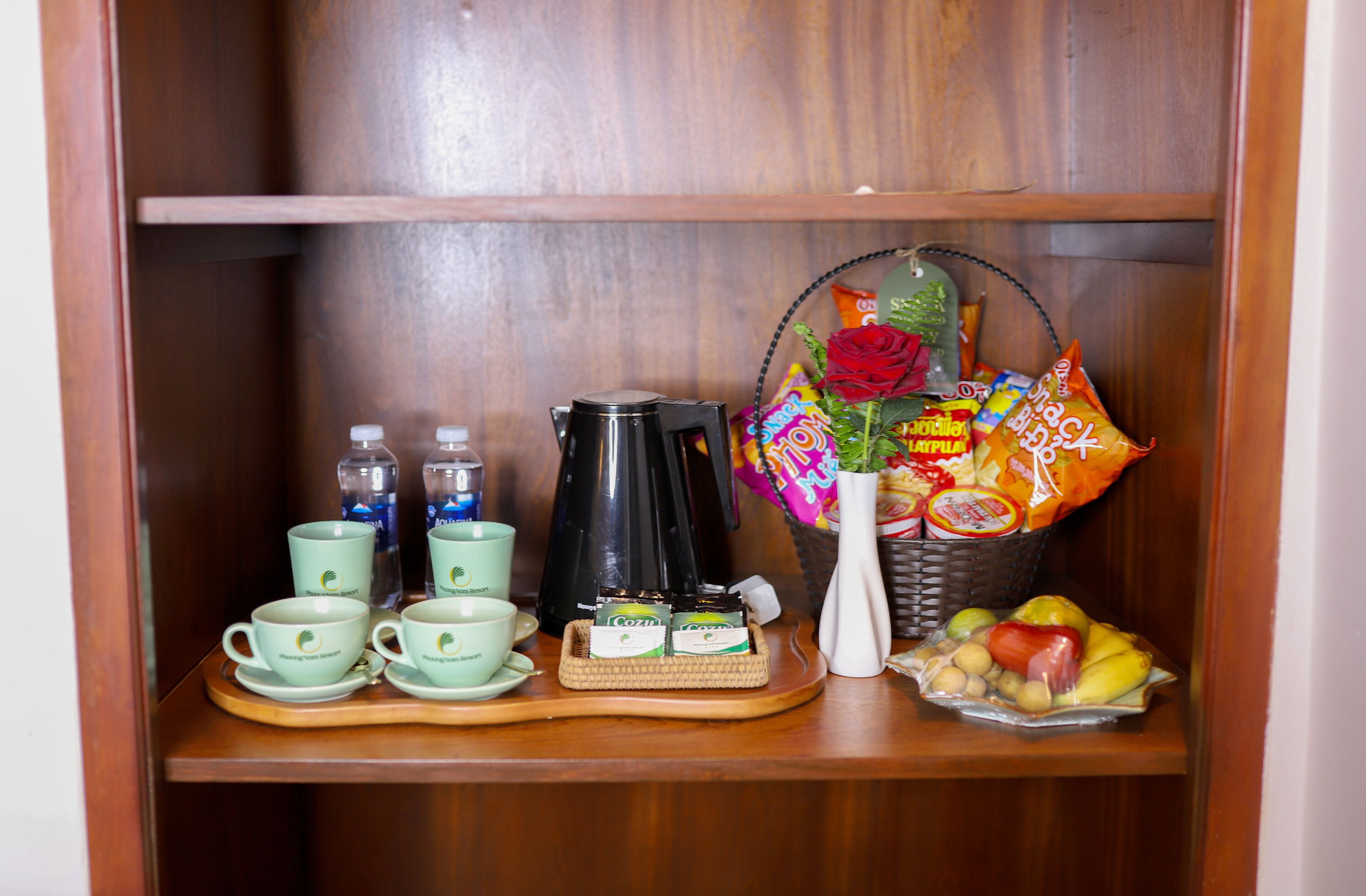 Una estantería de madera con una cafetera, tazas de té, botellas, una cesta de aperitivos y un florero con una rosa roja.