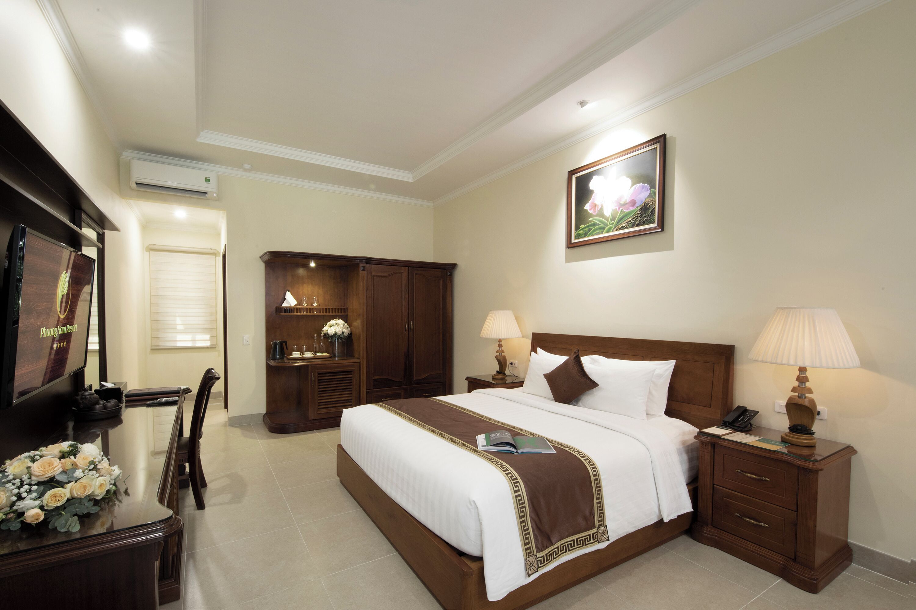 A hotel room with a large bed, a wooden nightstand with lamps, a desk with a TV, and a wardrobe.