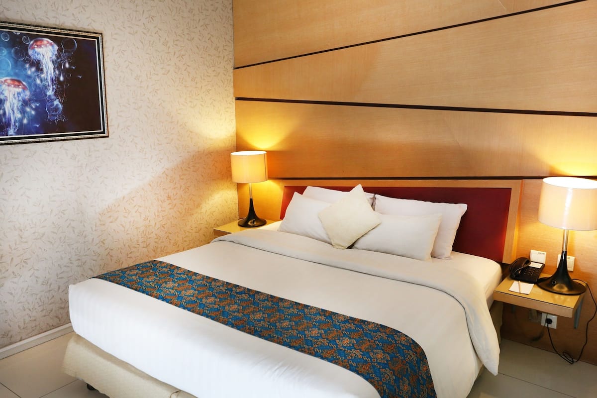 Quarto de hotel com uma cama grande, duas mesas de cabeceira, um encosto de cama vermelho, uma TV fixada na parede e um edredom estampado.