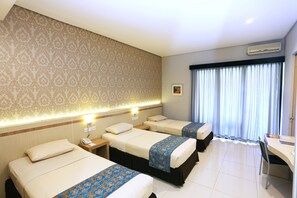 A hotel room with three beds, a desk, and a chair. There is a window with curtains, an air conditioning unit, and a patterned wallpaper on the wall. - Tanjung Kodok Beach Resort (Paciran)