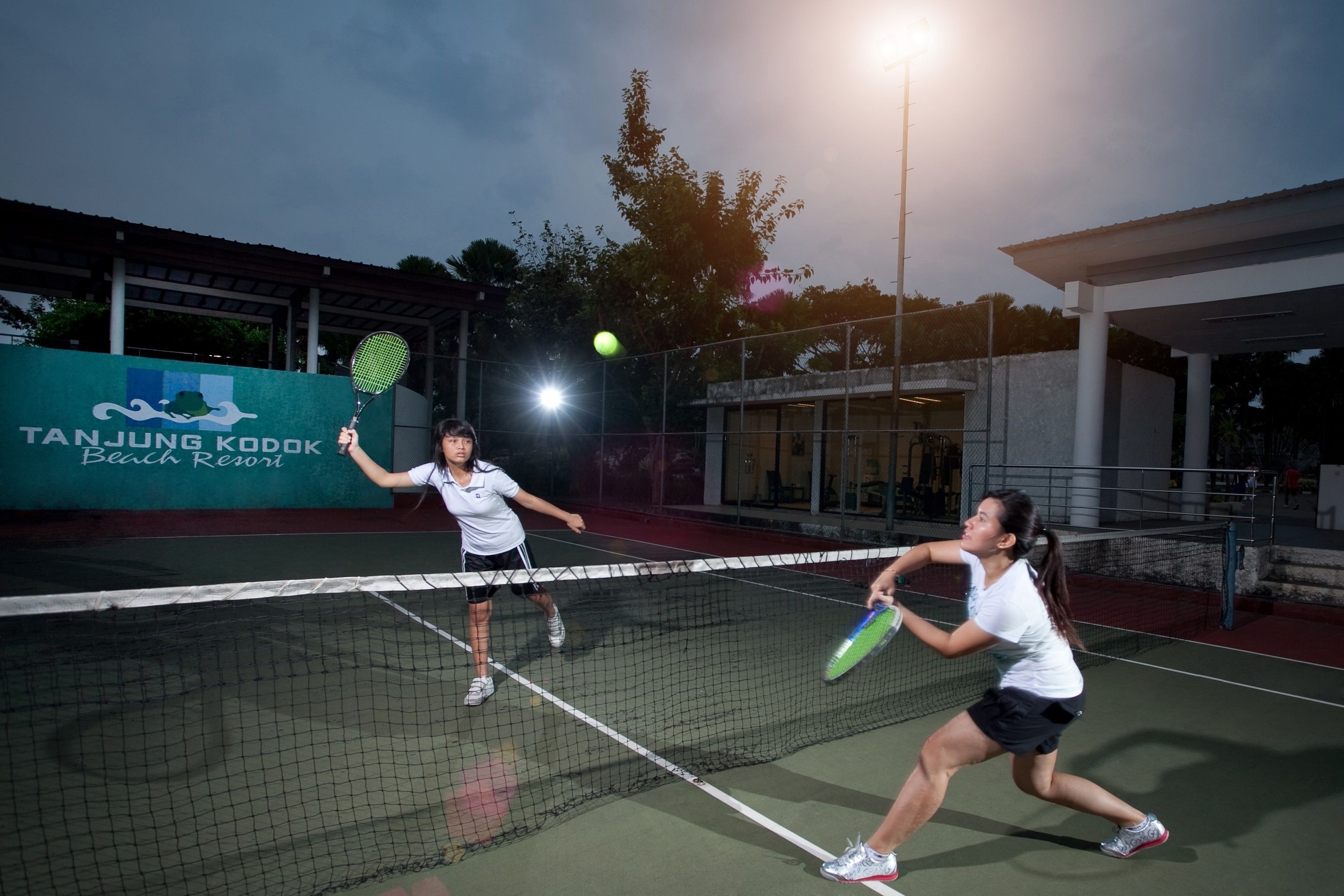A tennis court with a net in the middle, two players in white shirts and black shorts, and a banner in the background that reads "Tanjung Kordak Beach Resort".