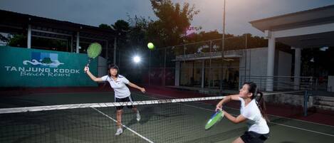 A tennis court with a net in the middle, two players in white shirts and black shorts, and a banner in the background that reads "Tanjung Kordak Beach Resort".