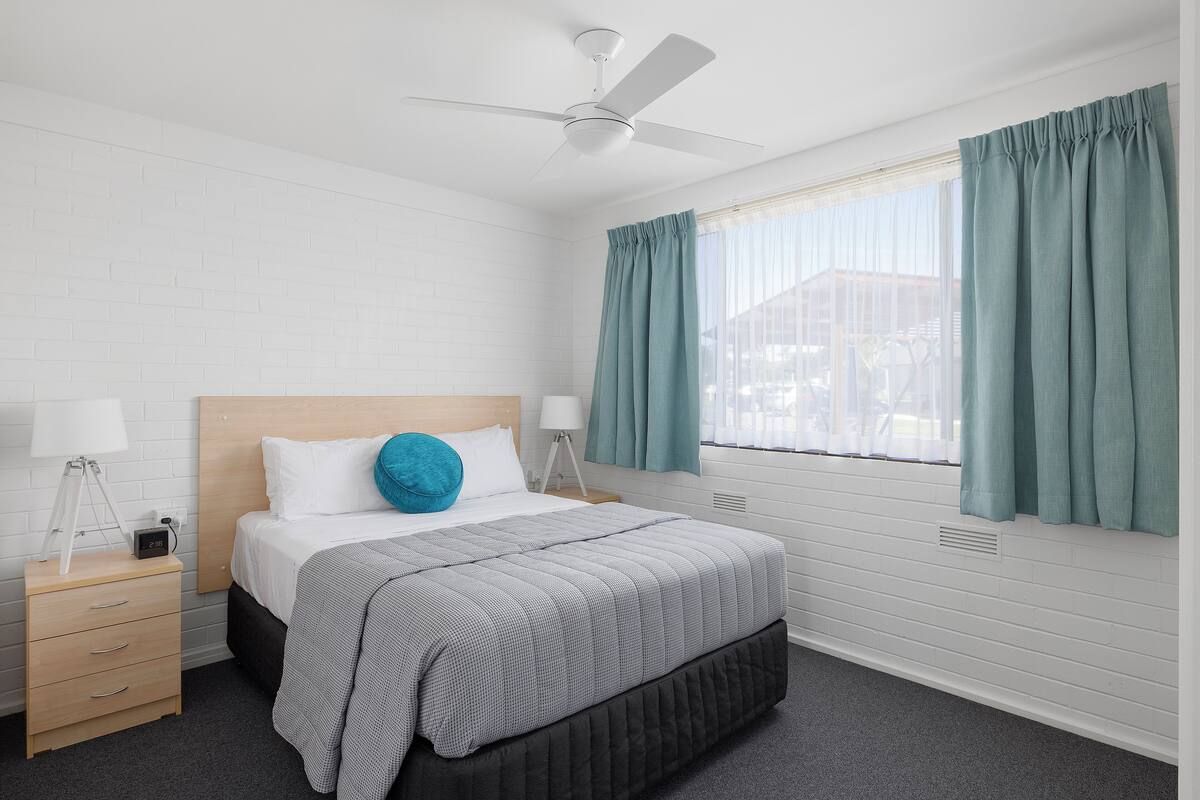 A bedroom with a bed, a nightstand, a lamp, a ceiling fan, and a window with curtains.