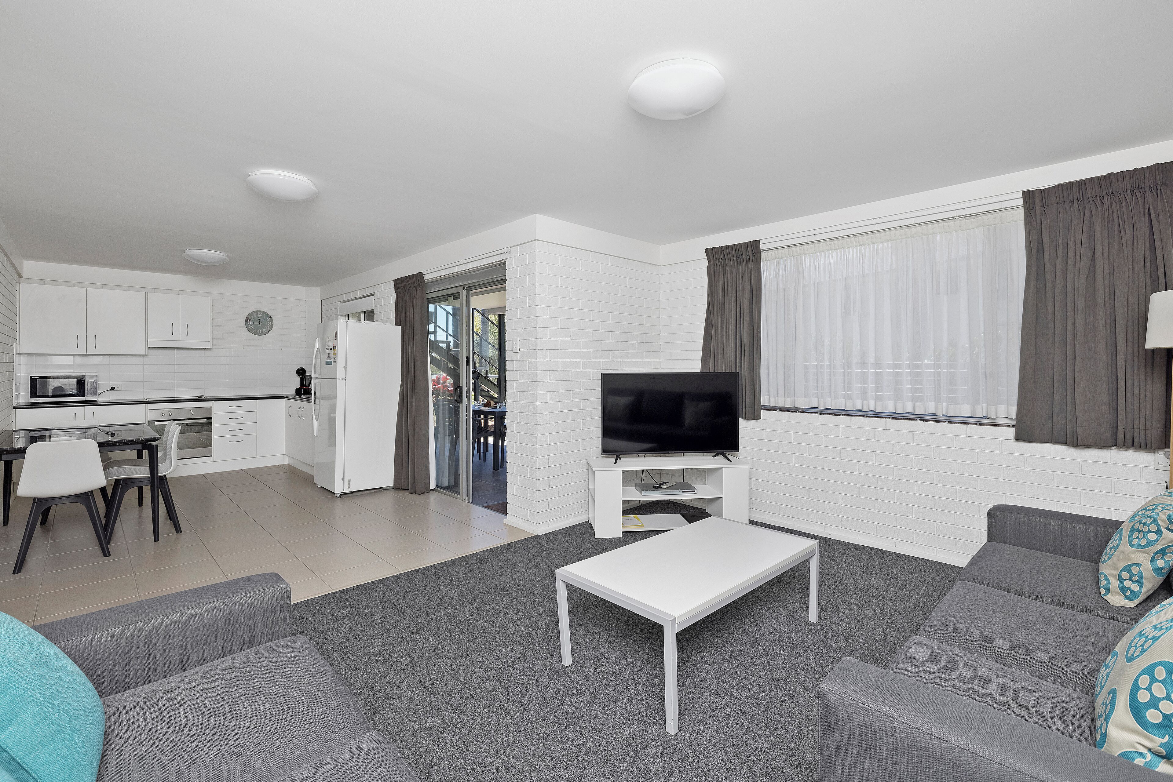 Een moderne woonkamer met een grijze bank, een witte salontafel en een televisie op een witte dressoir. Er is een zithoek met een tafel en stoelen, en een keuken met witte kasten en een koelkast.