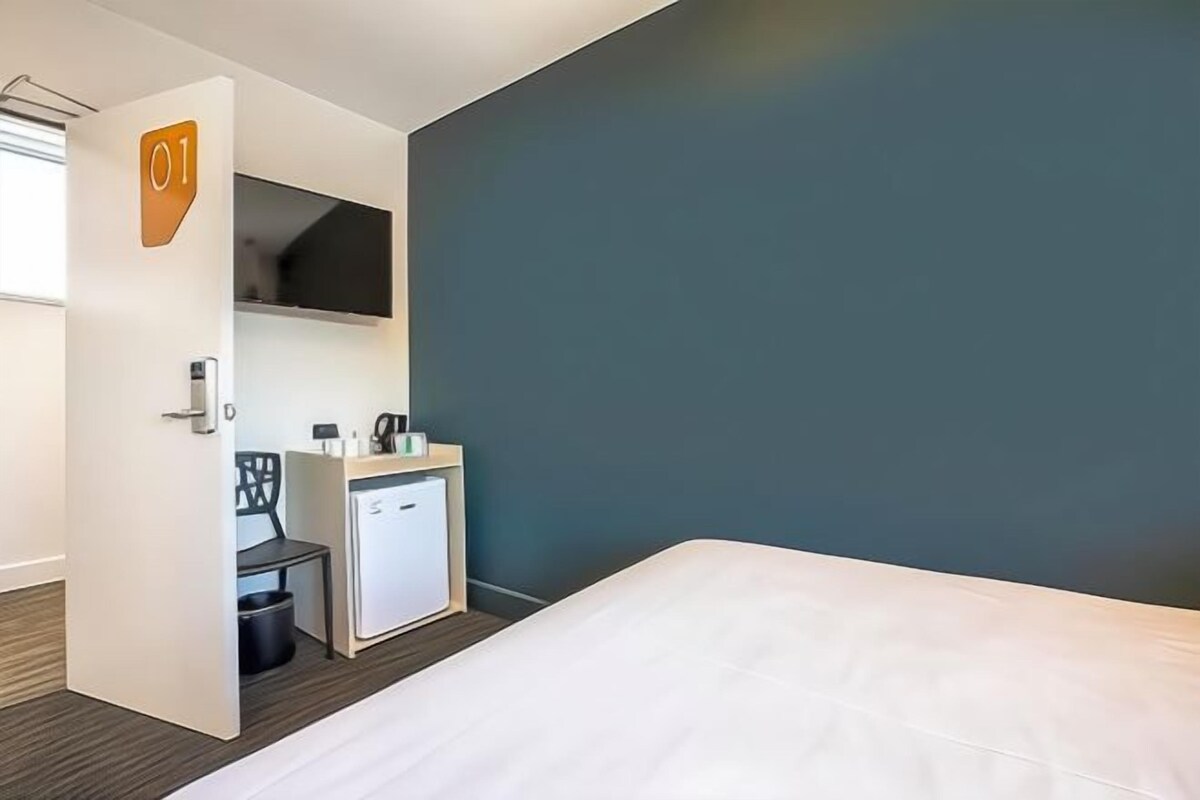 A hotel room with a bed, a desk with a chair, a small refrigerator, and a television on the wall.