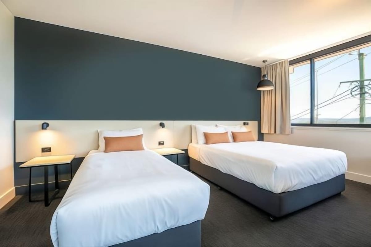 A hotel room with two beds, a large window with a view, and a dark-colored wall.